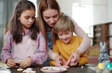Lady with two children, leaning over one child helping them bake cookies